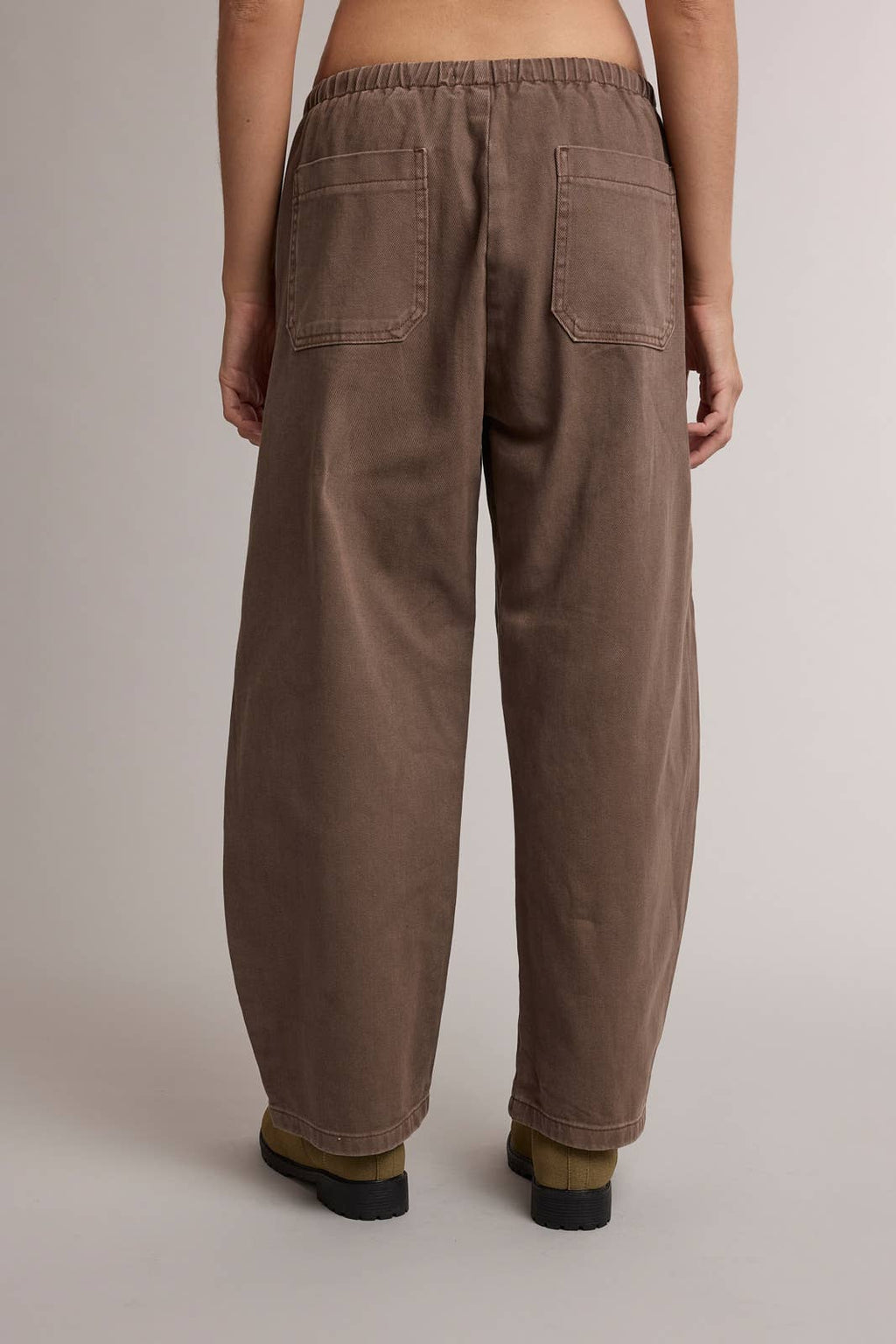 A person shown from the back wearing loose, wide-leg brown pants with an elastic waistband and two large back patch pockets, photographed against a plain light gray studio background.