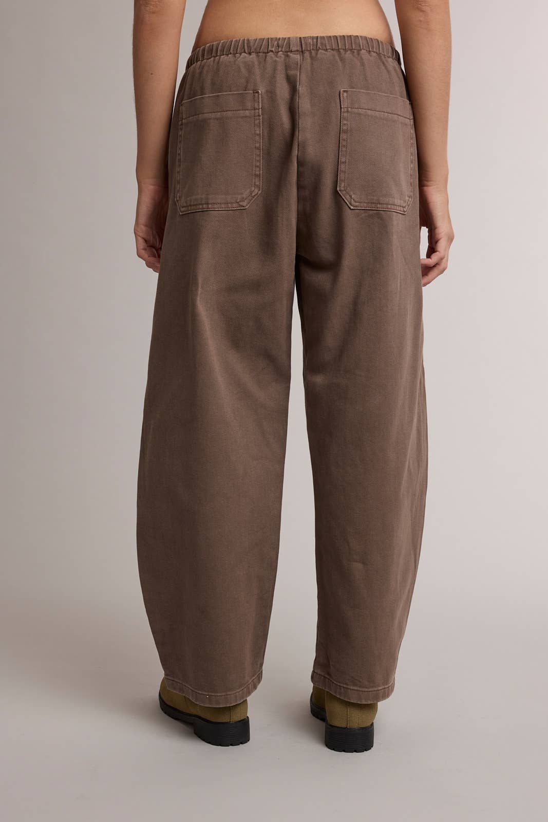A person shown from the back wearing loose, wide-leg brown pants with an elastic waistband and two large back patch pockets, photographed against a plain light gray studio background.