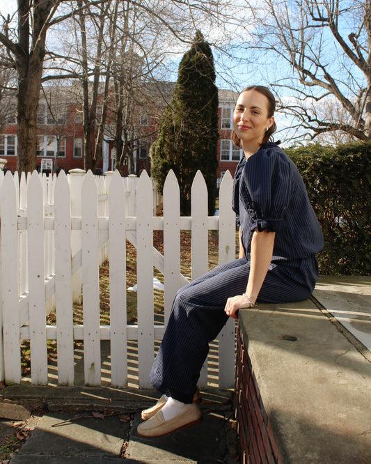Woman sitting on a white picket fence with bare trees and a clear sky in the background
