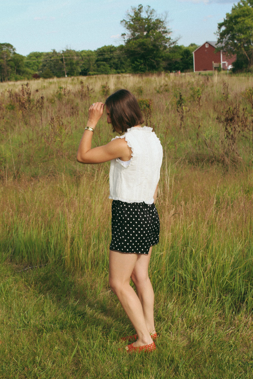 Woman standing in a field wearing a white sleeveless top and black polka dot shorts.