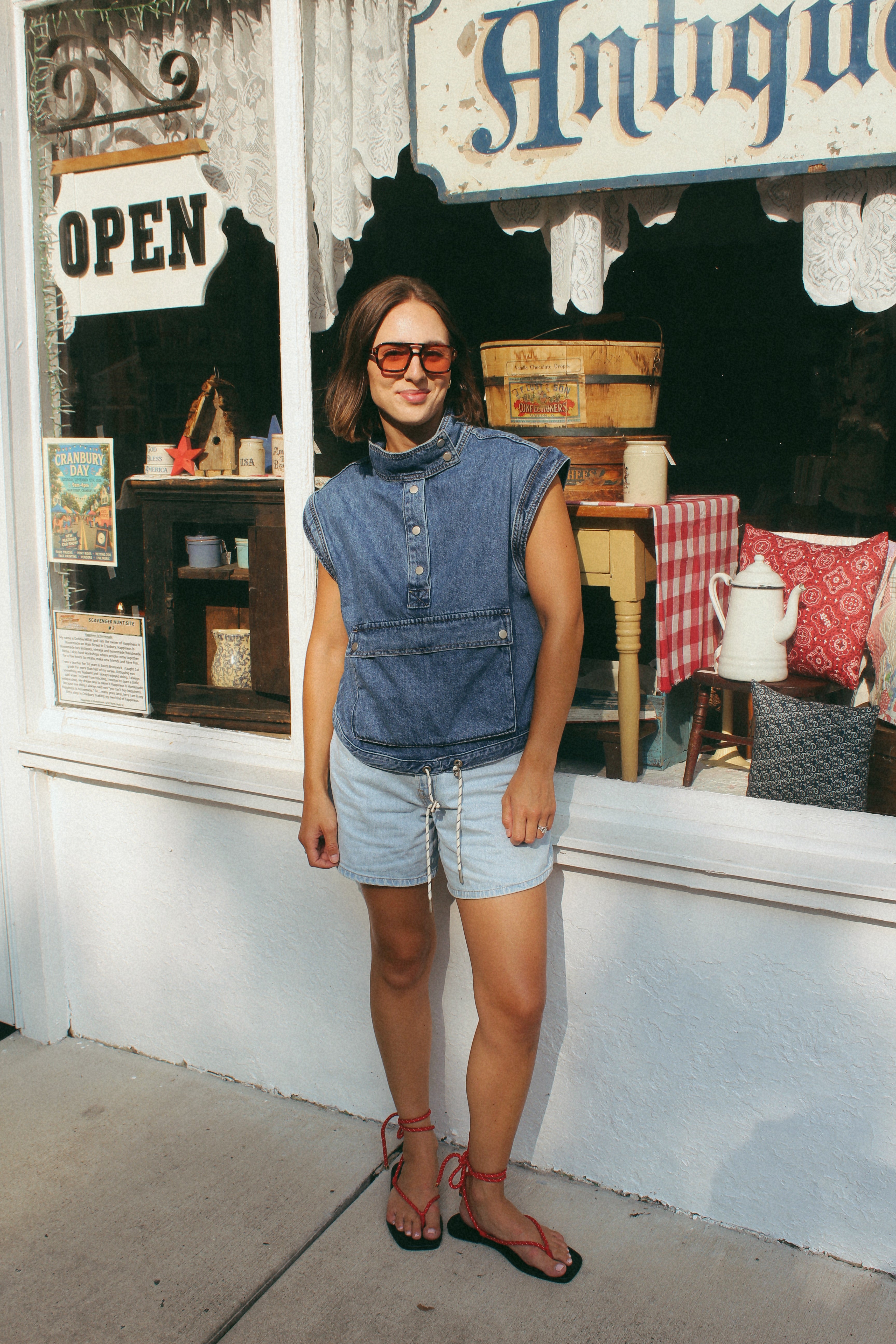 A woman wearing a sleeveless denim top and light denim shorts stands in front of an antique shop window.