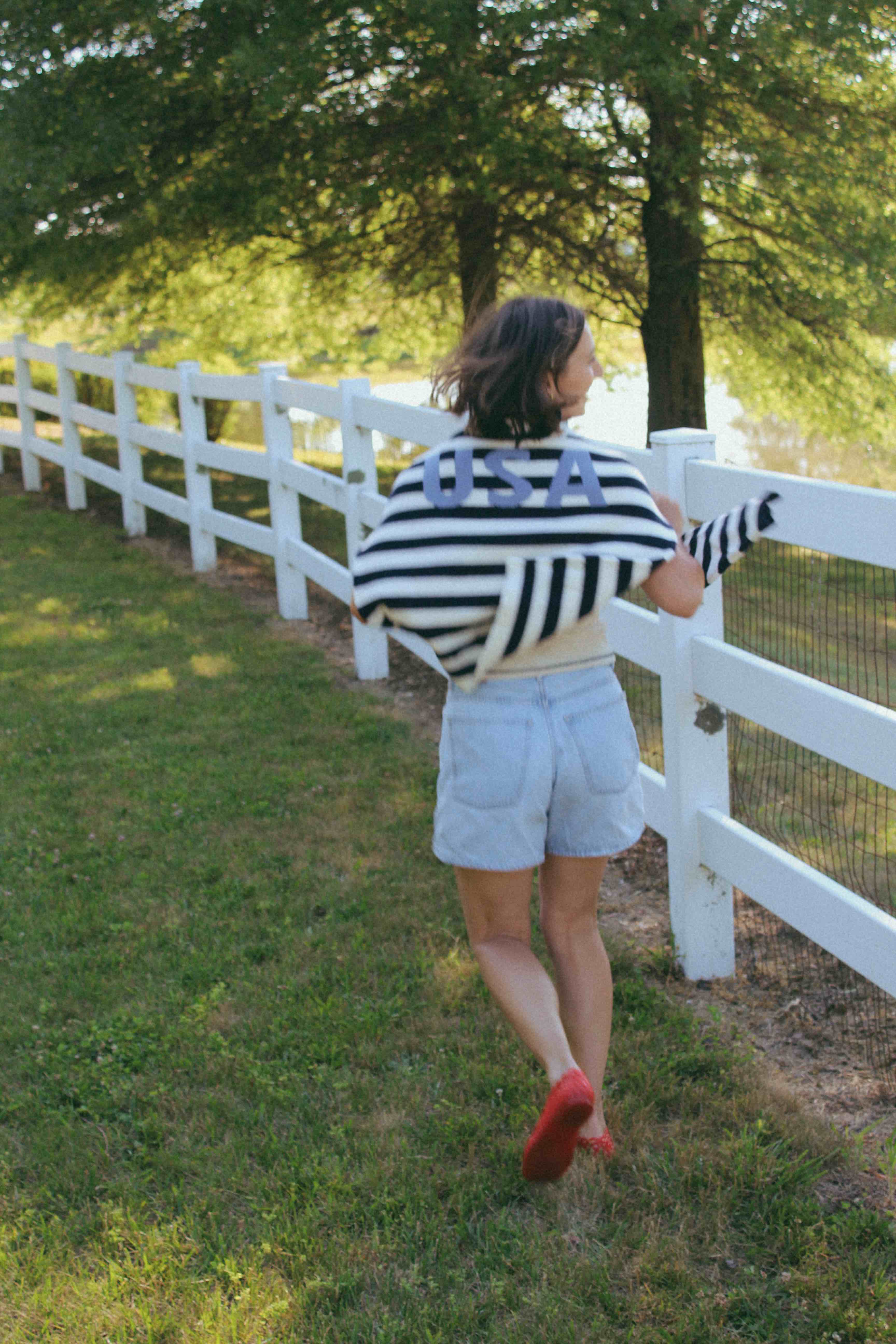 Person wearing a striped shirt with 'USA' on it, running towards a white fence in a park-like setting.