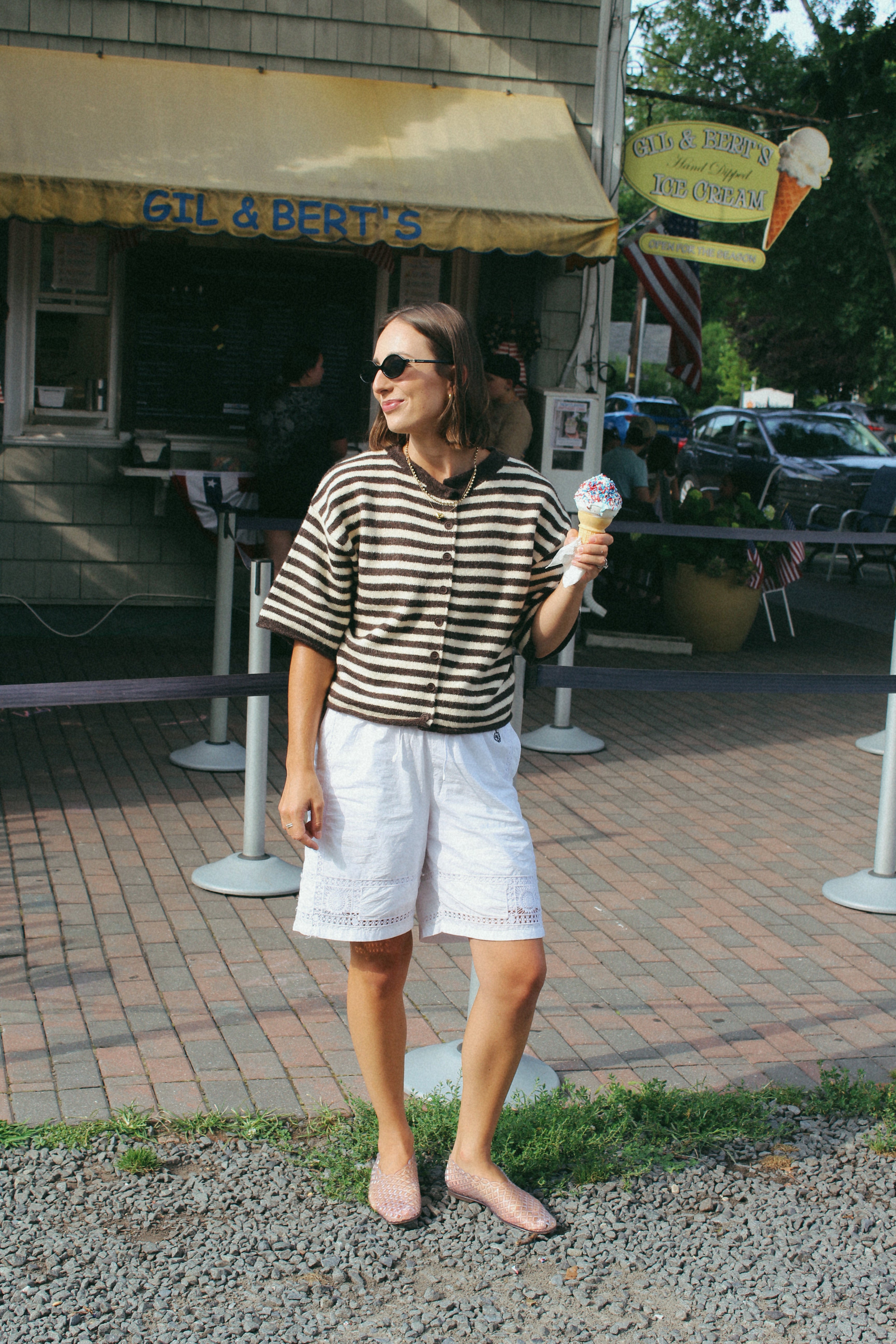 A woman stands outdoors in front of a restaurant, wearing a black and beige horizontally striped short-sleeve button-up shirt, white shorts, and sunglasses while holding an ice cream cone.