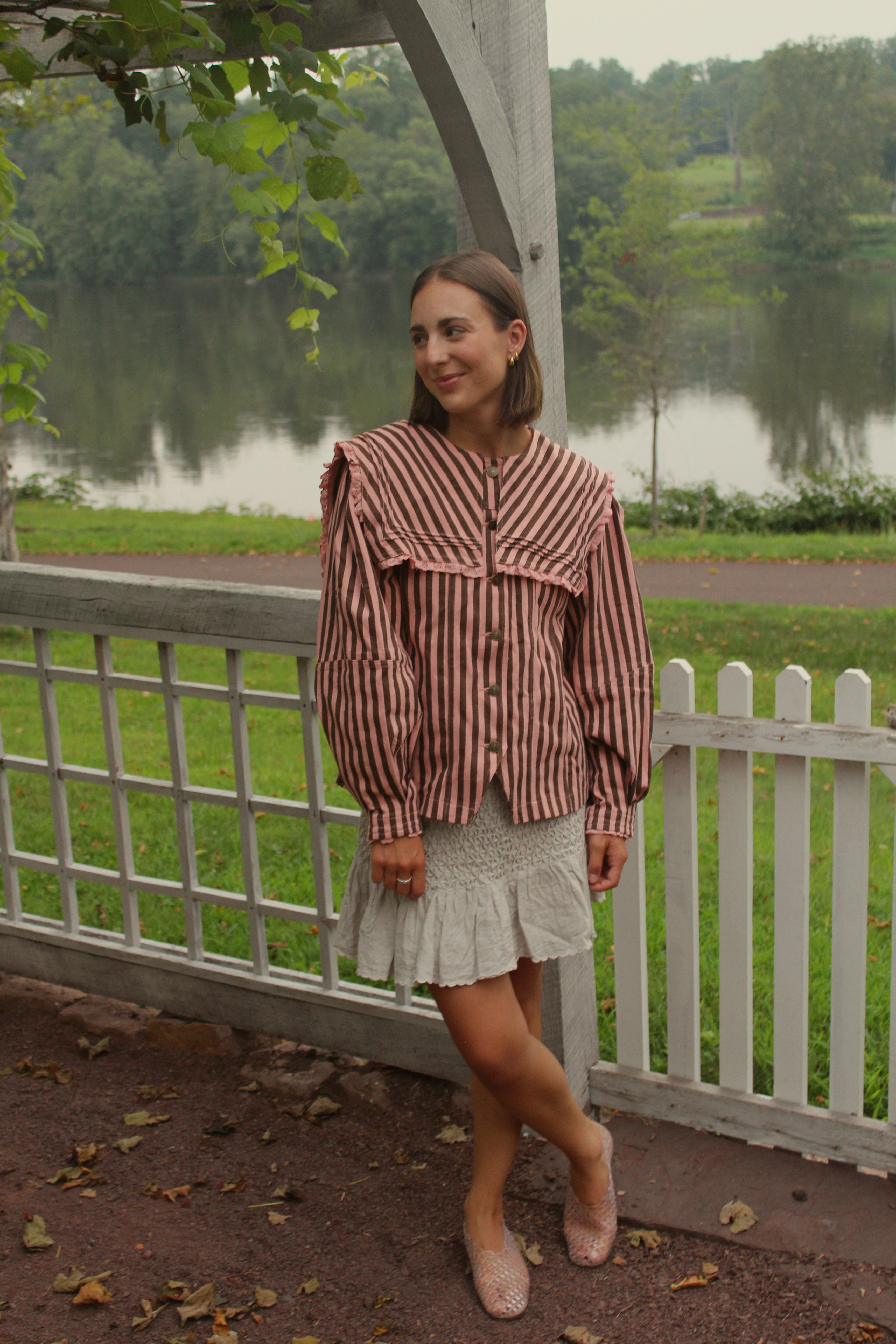 A woman stands outdoors by a white picket fence next to a lake, wearing a brown and pink vertically striped ruffled blouse, a light gray ruffled mini skirt, and beige pointed flats.