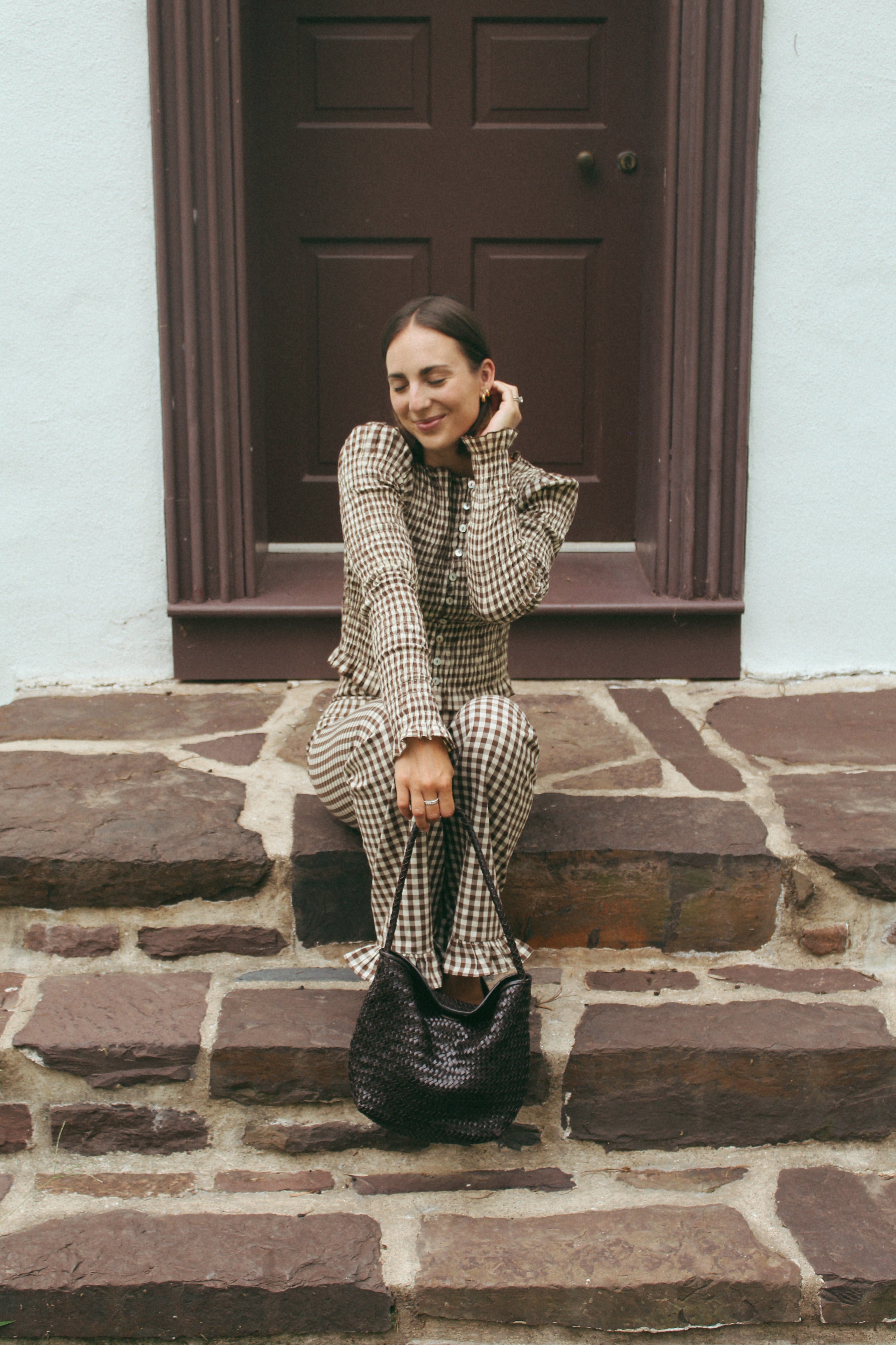 A woman wearing a beige and black checkered long-sleeve top and matching trousers sits on stone steps in front of a dark brown door, holding a black textured handbag.