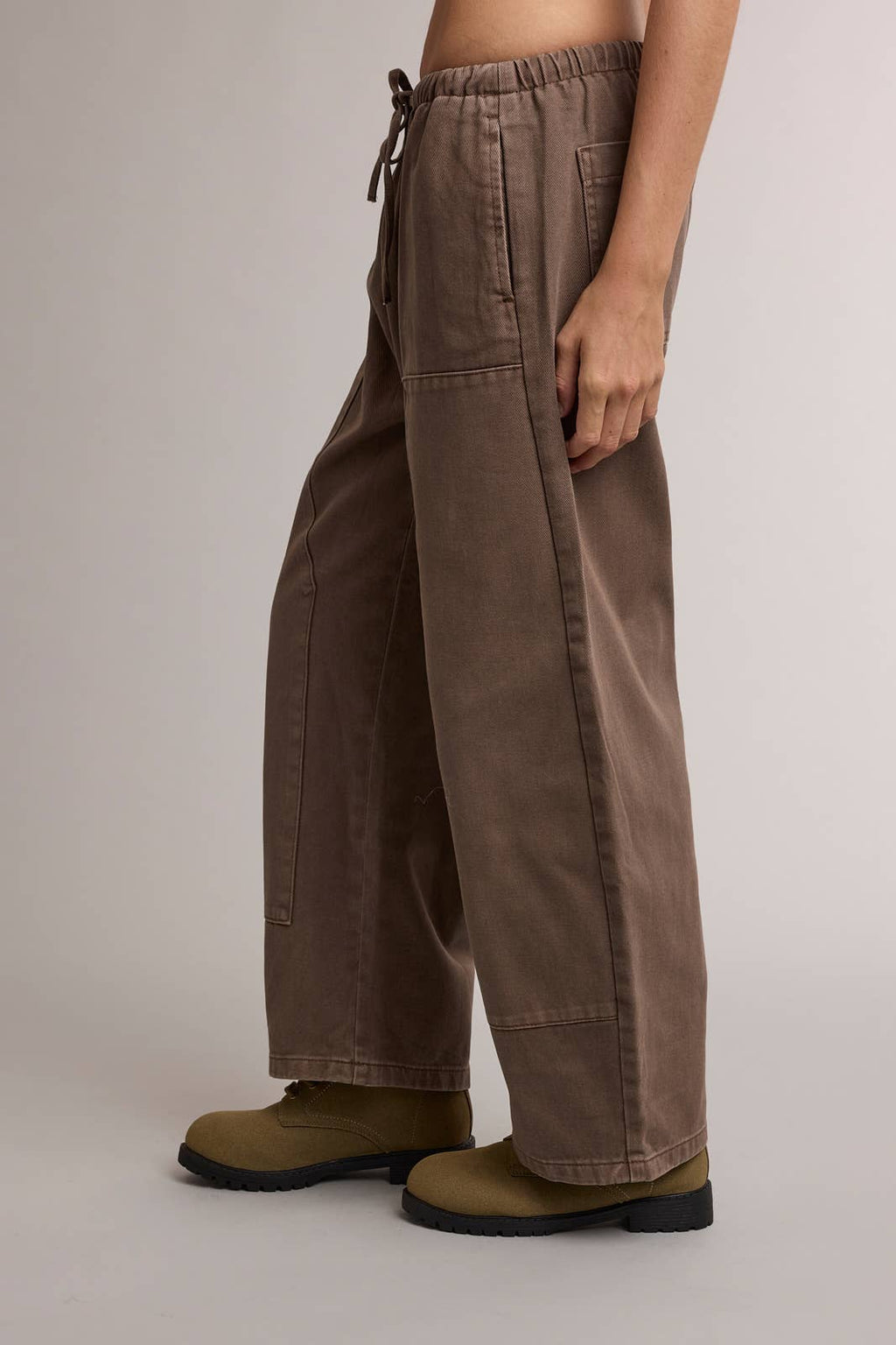 A person wearing wide-leg brown pants with a drawstring waist and olive green work boots, shown from the waist down against a plain light gray studio background.