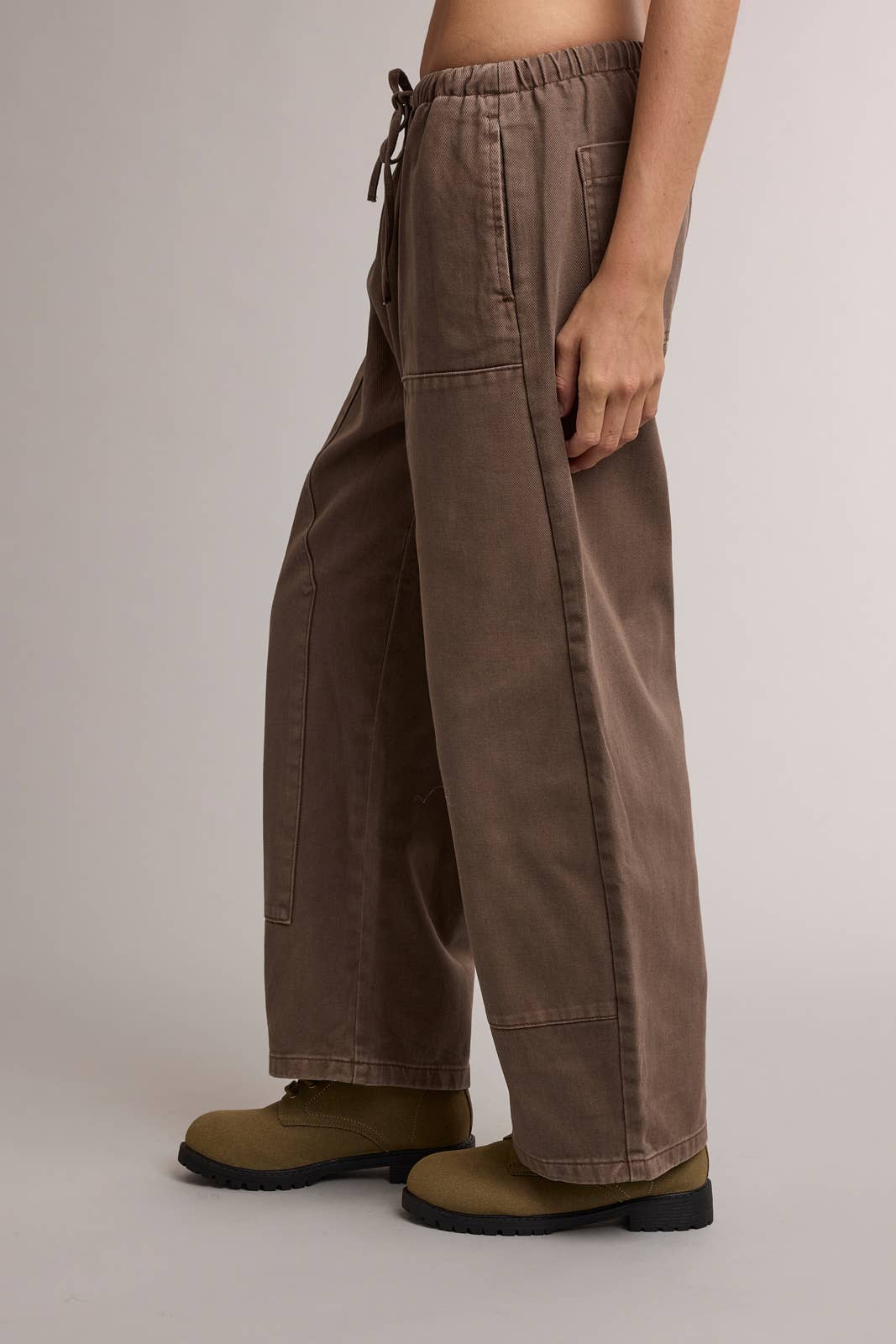 A person wearing wide-leg brown pants with a drawstring waist and olive green work boots, shown from the waist down against a plain light gray studio background.